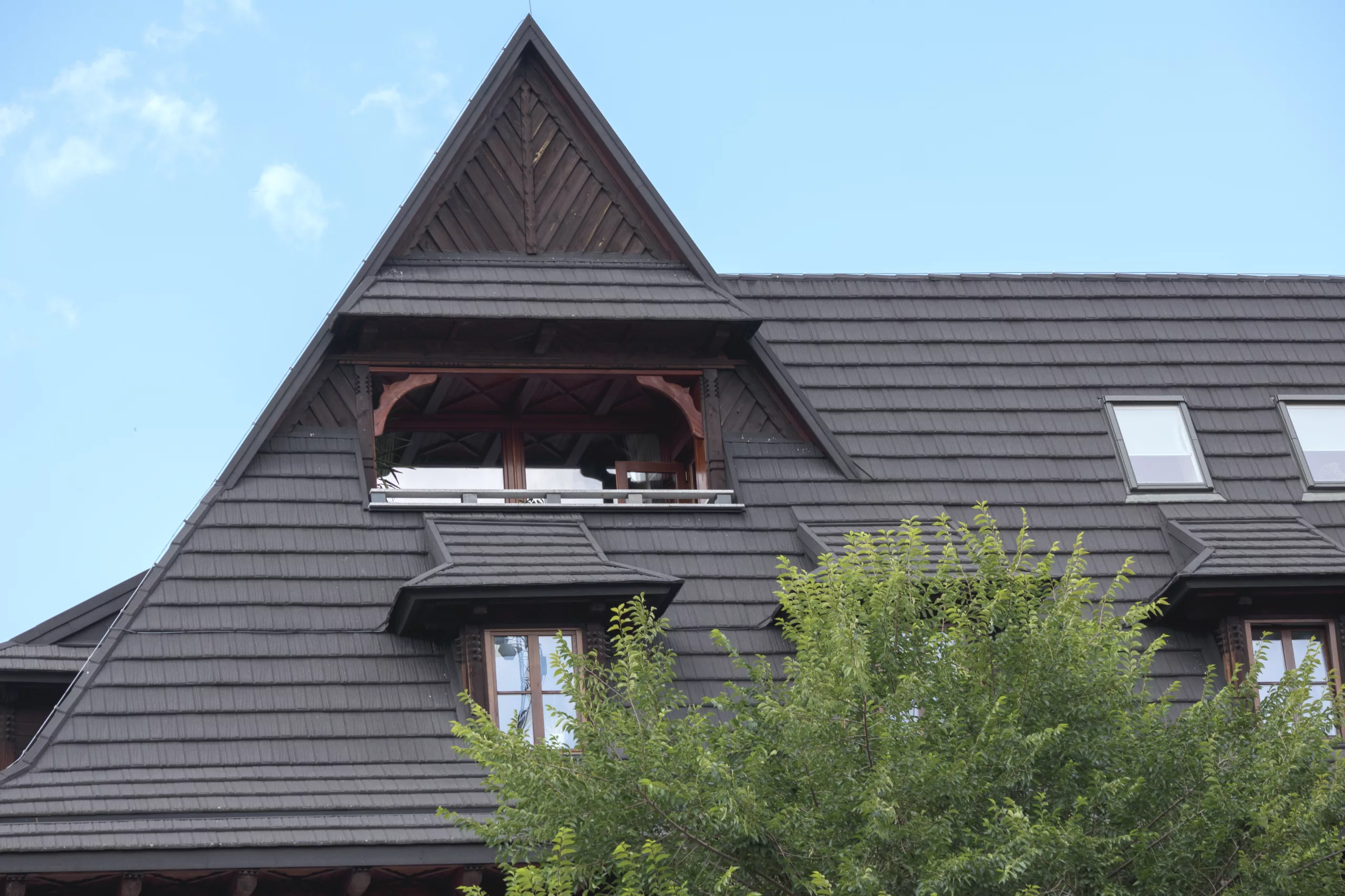 The image shows the upper portion of a house with a beautiful brown roof and a peaked roof on one side. There is a large tree near the house